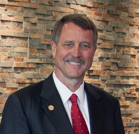 Smiling, Caucasian male with a mustache wearing a blue suit, white collared shirt, red tie with print, gold lapel pin, standing against a brick wall, posing for a photo