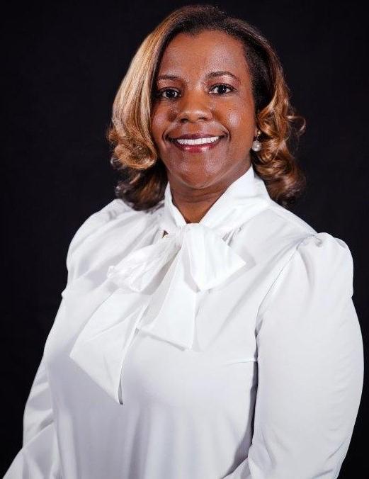 An African American female, Dr. Kristie Searcy, Dean of Health Sciences, has medium length blonde curly hair and smiles wearing a white dress shirt with a bow neck and pearl earrings against a black backdrop.